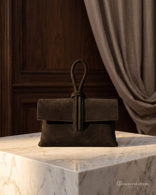 La pochette Capri en cuir suède noir posé sur un bloc de marbre blanc en boutique