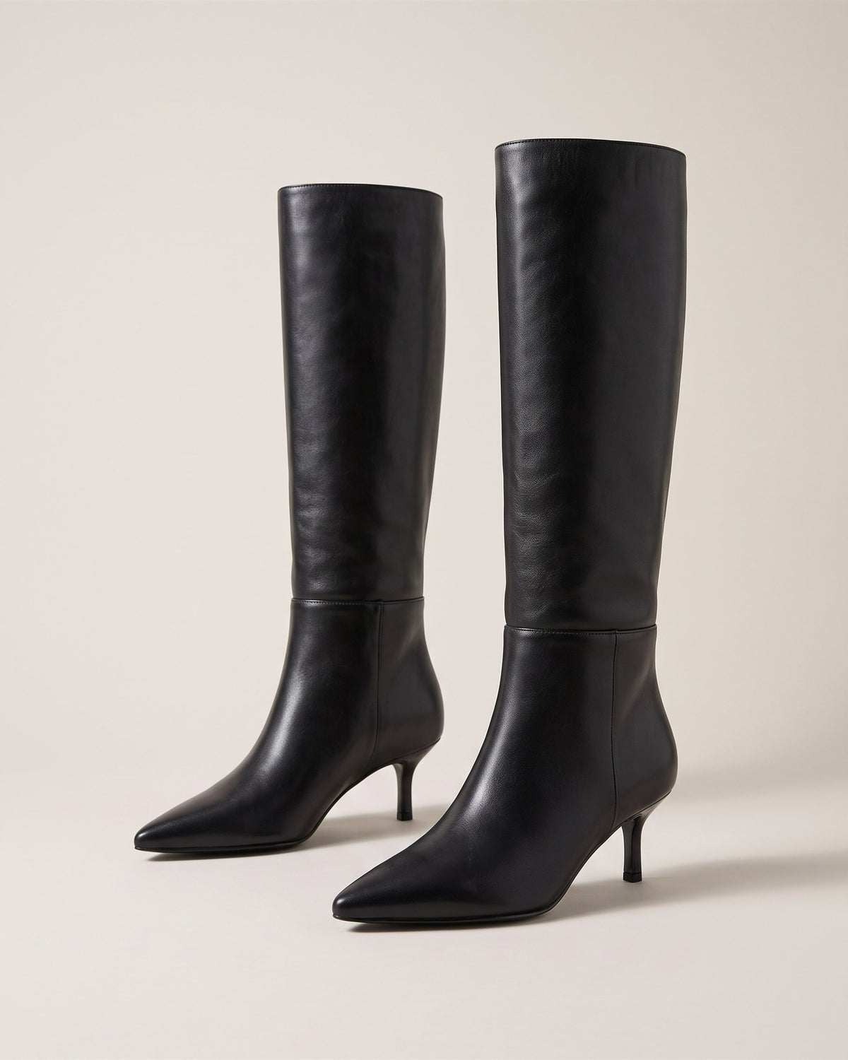 Bottes hautes noires à talon fin et bout pointu, vue de face sur fond pierre