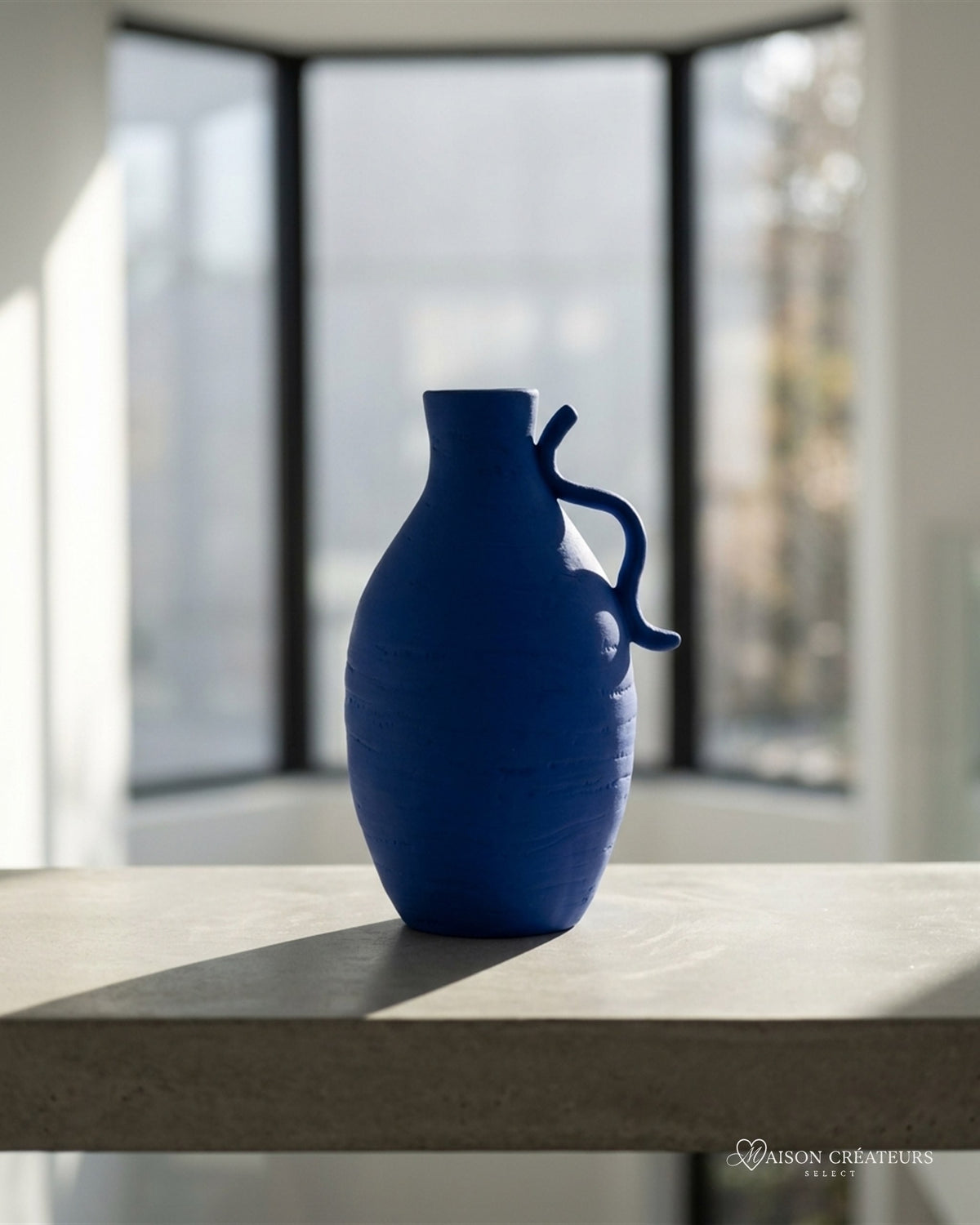 Vase Creta en céramique bleu Klein à anse, posé sur du béton dans un atrium lumineux