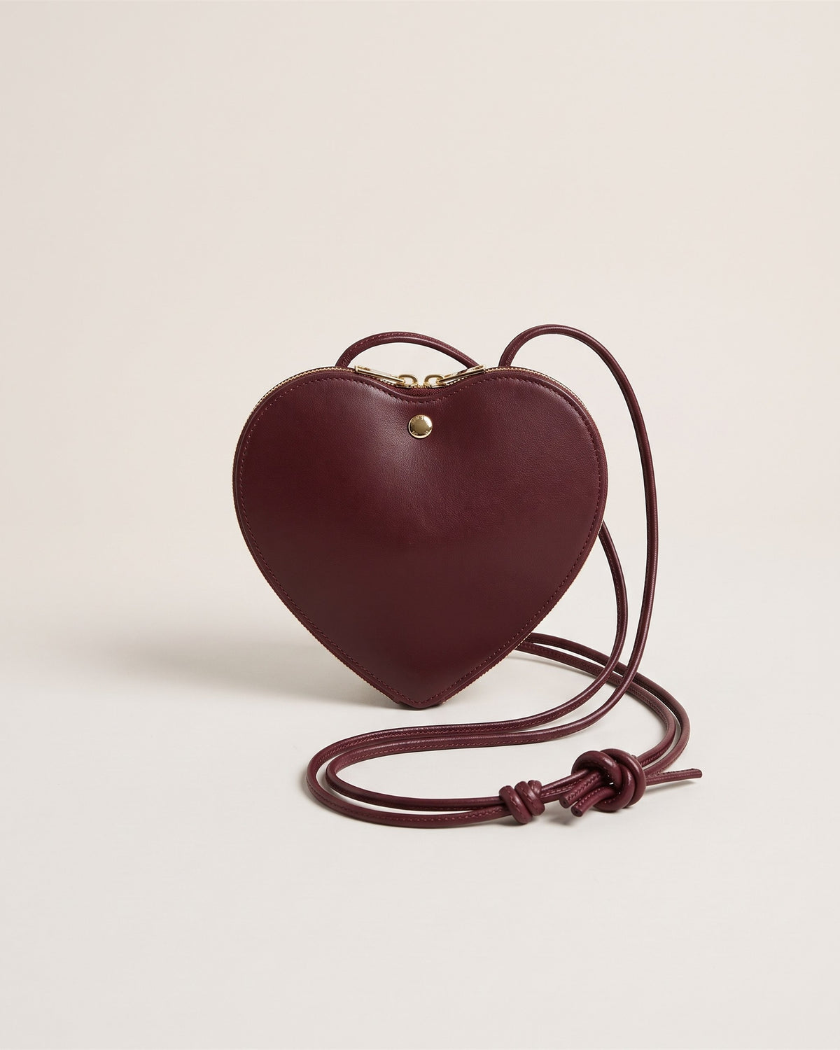 La pochette en forme de cœur en cuir lisse bordeaux, avec sa fine bandoulière en cuir nouée, vue de face sur fond pierre