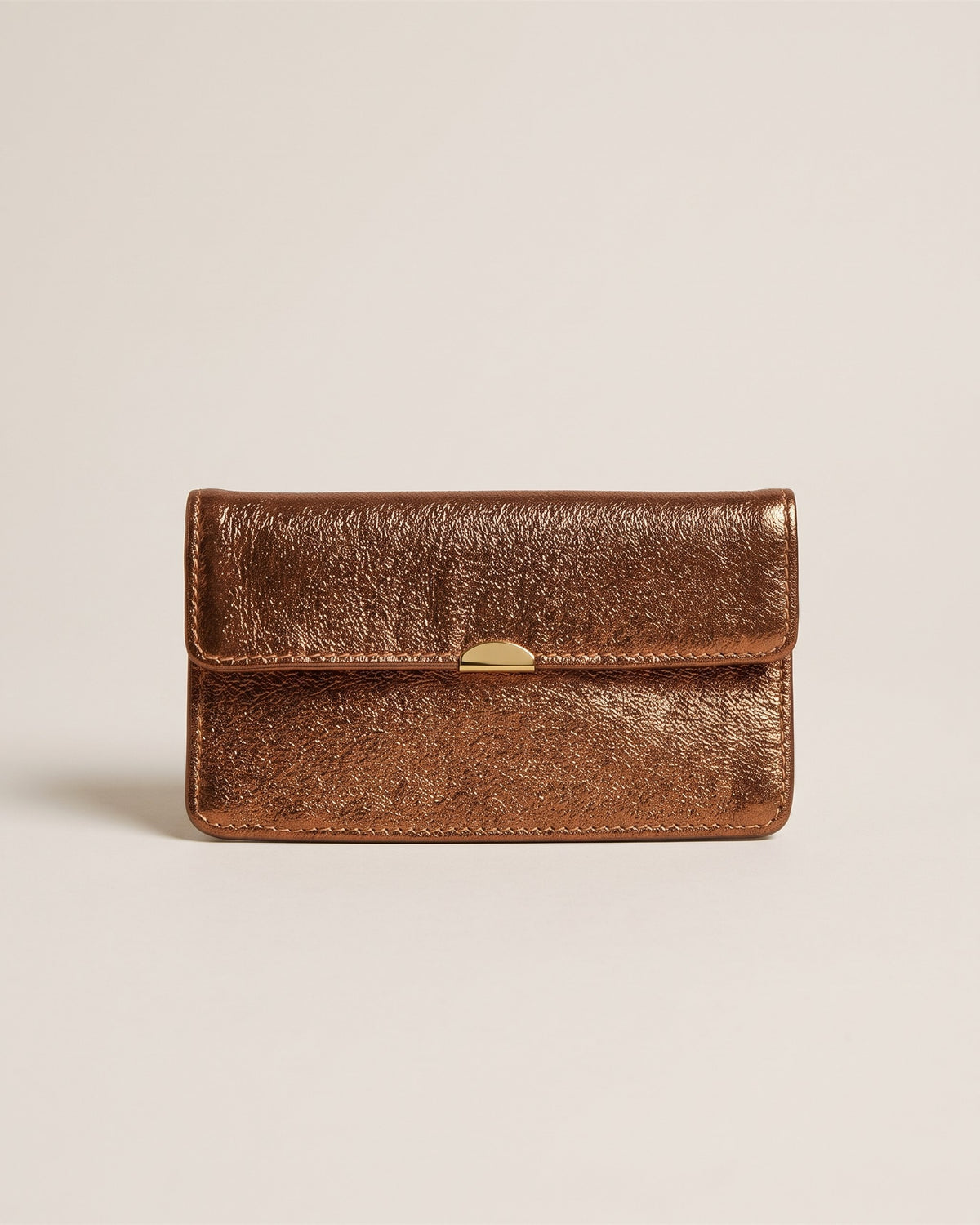 Le porte-monnaie compact tout-en-un en cuir métallisé noisette, forme enveloppe à rabat et fermoir doré, vue de face sur fond pierre