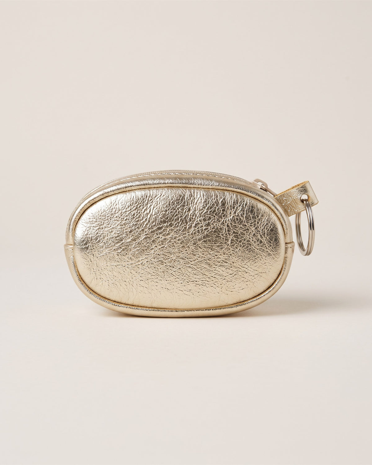 Le porte-monnaie porte-clés en cuir métallisé doré, forme ovale zippée avec anneau porte-clés, vue de face sur fond pierre