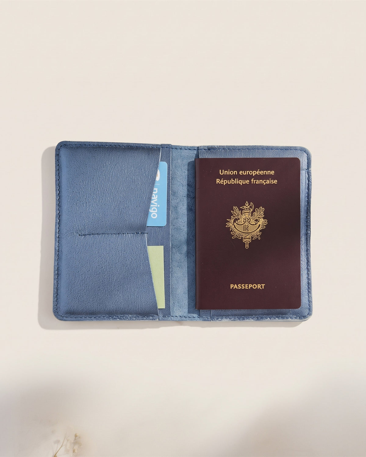 Le porte-passeport en cuir bleu marine, ouvert avec un passeport français glissé dans sa poche, vue de face sur fond pierre