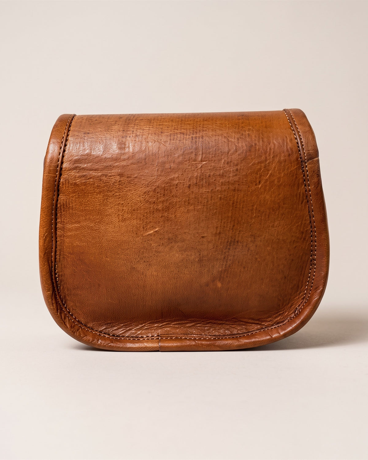 Le sac bandoulière en cuir cognac vieilli 18 cm, forme besace arrondie avec rabat et coutures apparentes, vue de face sur fond pierre