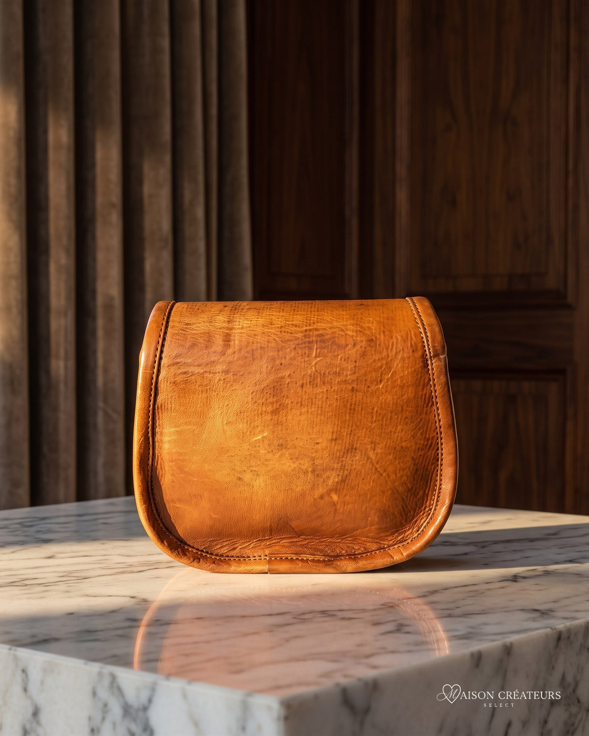 Le sac bandoulière en cuir cognac vieilli 18 cm posé sur un bloc de marbre blanc en boutique