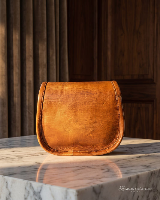 Le sac bandoulière en cuir cognac vieilli 18 cm posé sur un bloc de marbre blanc en boutique