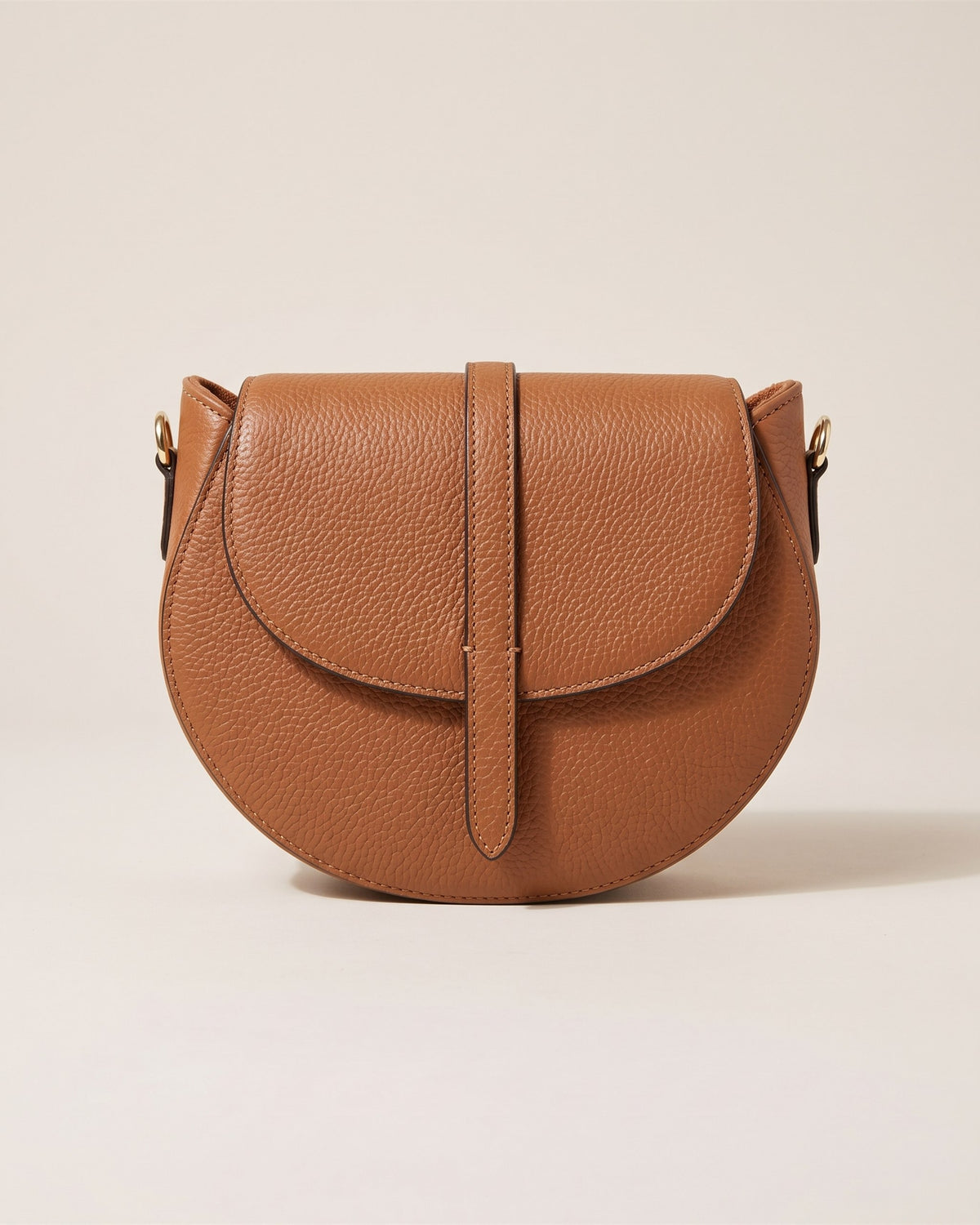Le sac bandoulière Madelena en cuir grainé camel, forme demi-lune, rabat à languette et bandoulière fine, vue de face sur fond pierre