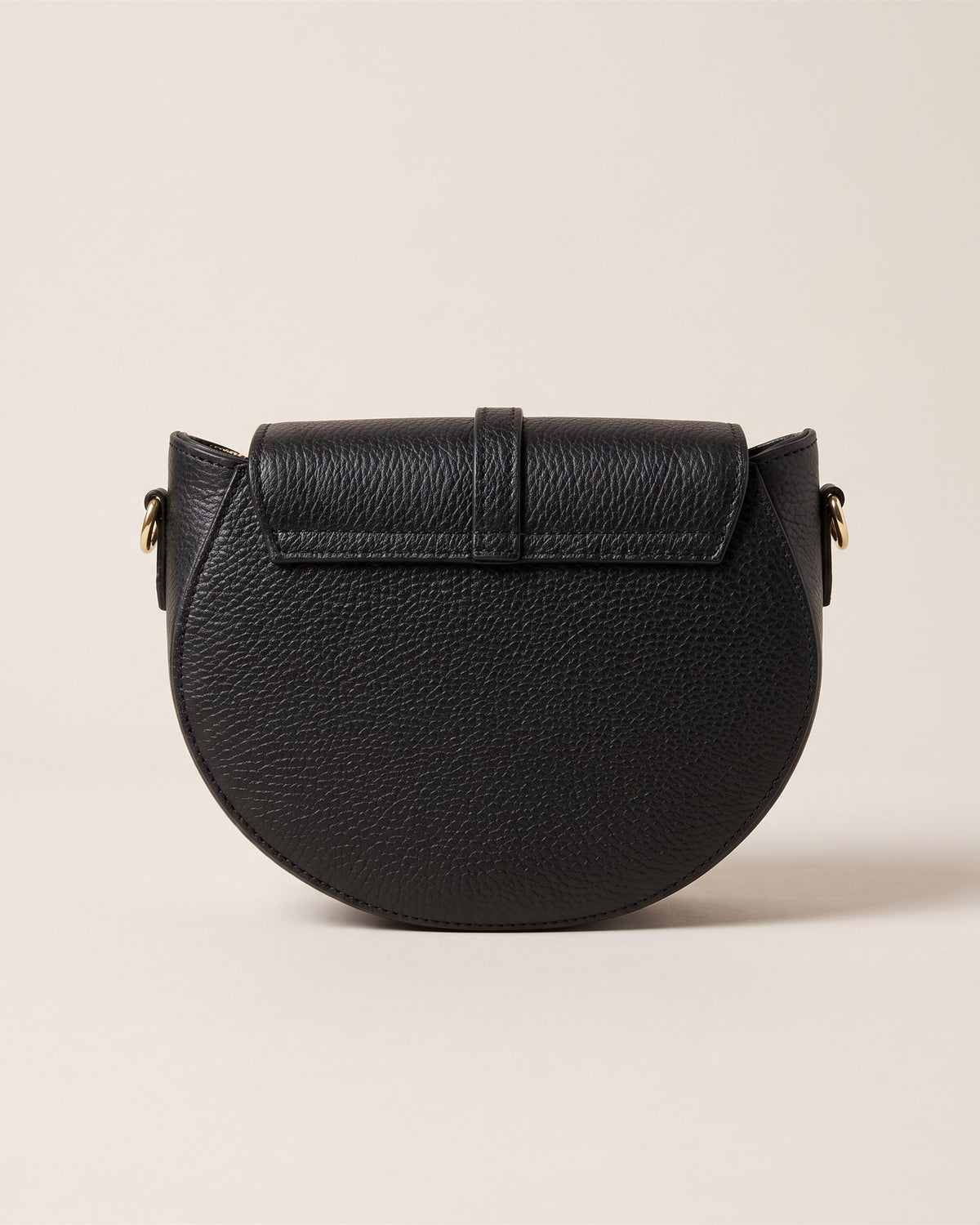 Le sac bandoulière Madelena en cuir grainé noir, forme demi-lune, rabat à languette et bandoulière fine, vue de face sur fond pierre