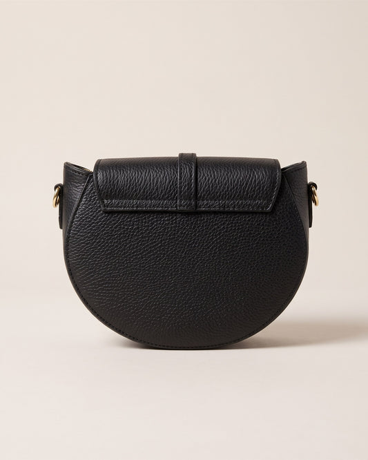 Le sac bandoulière Madelena en cuir grainé noir, forme demi-lune, rabat à languette et bandoulière fine, vue de face sur fond pierre