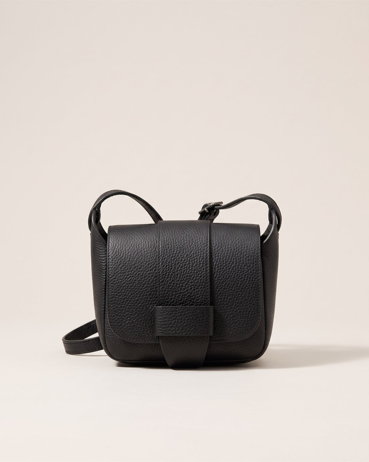 Le sac bandoulière Margot en cuir grainé noir, forme arrondie, rabat à bouton pression et bandoulière fine, vue de face sur fond pierre