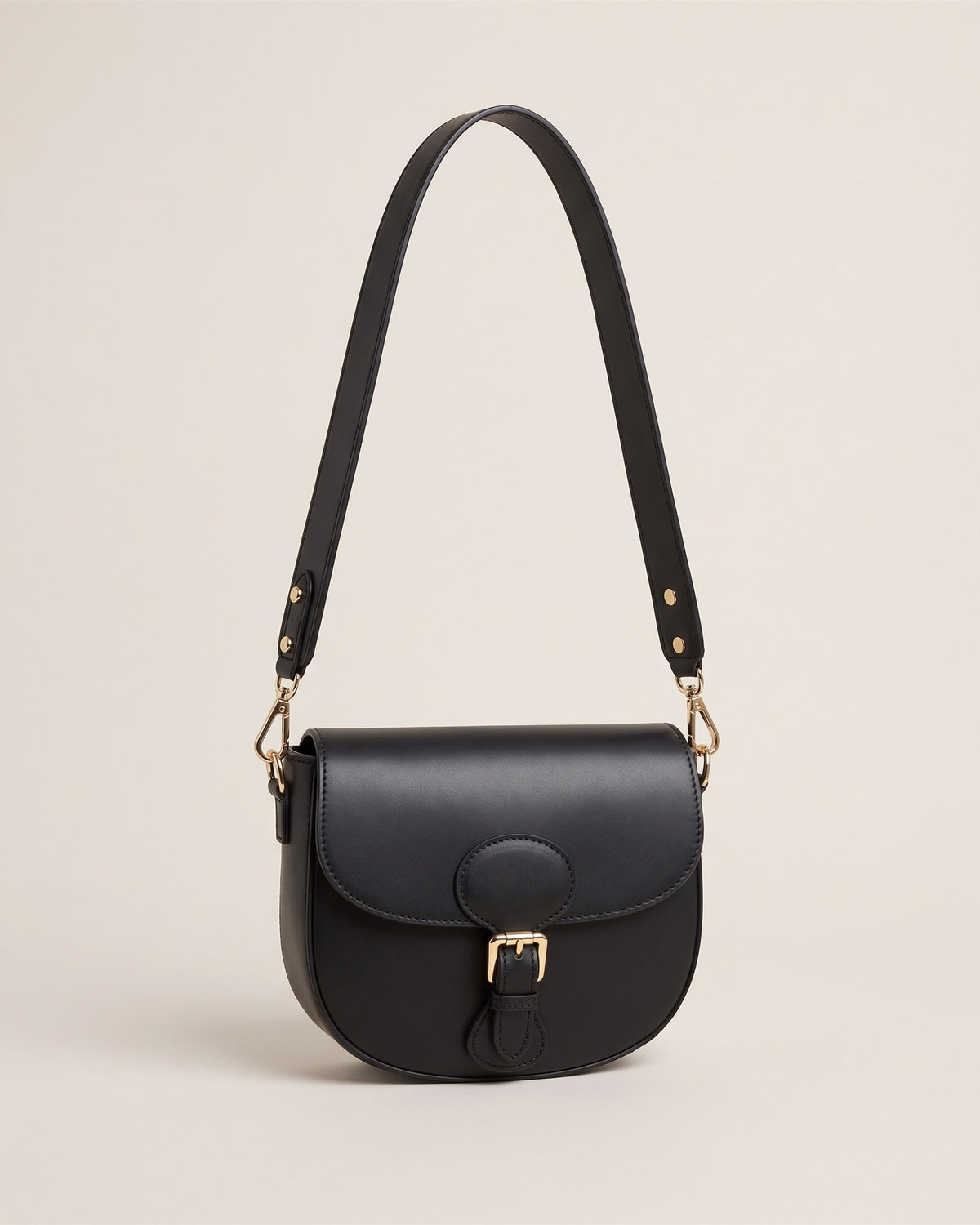 Le sac bandoulière Leonine en cuir lisse noir, forme besace à rabat avec boucle dorée et bandoulière large, vue de face sur fond pierre