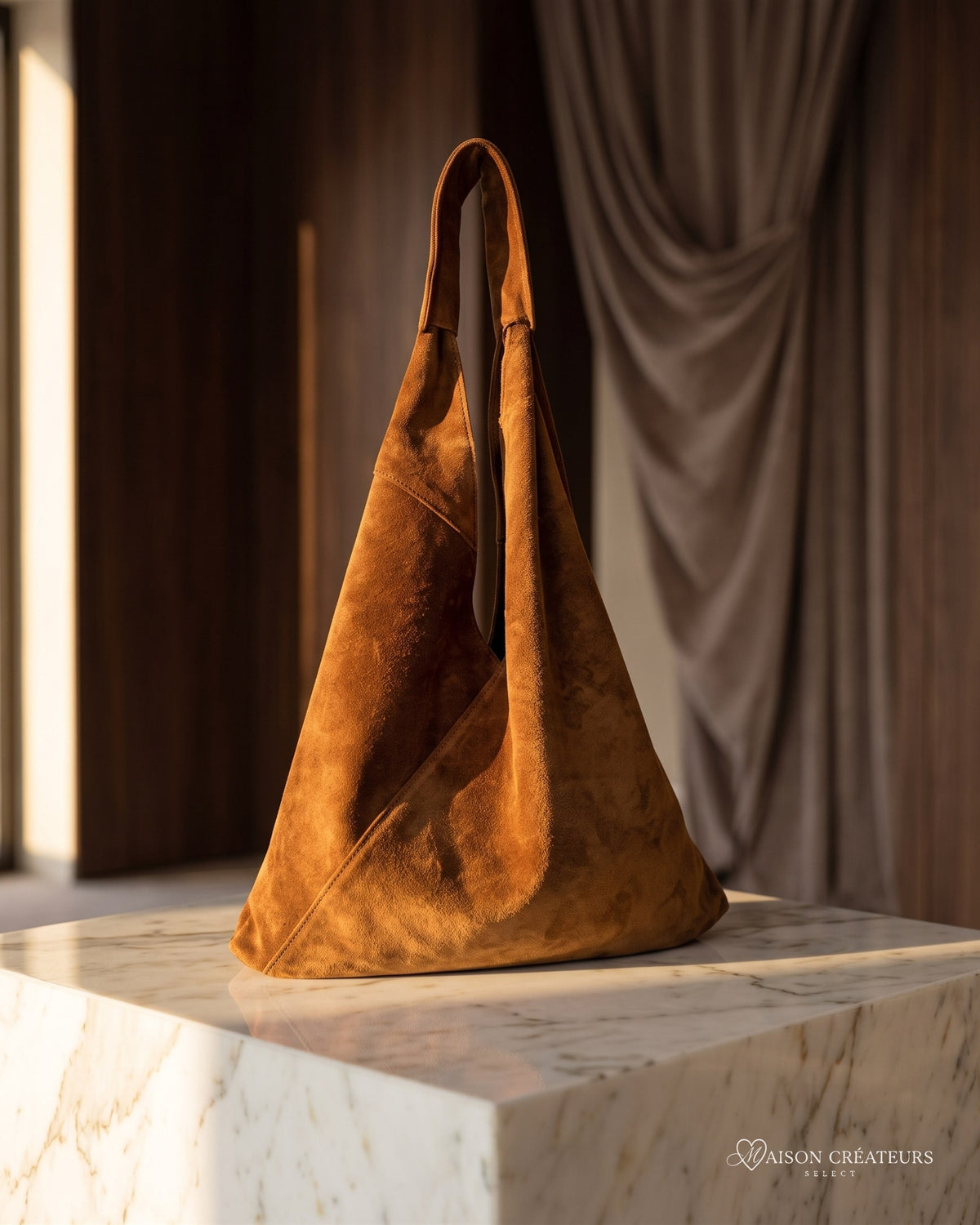 Le sac Nomade en cuir suède camel posé sur un bloc de marbre blanc en boutique