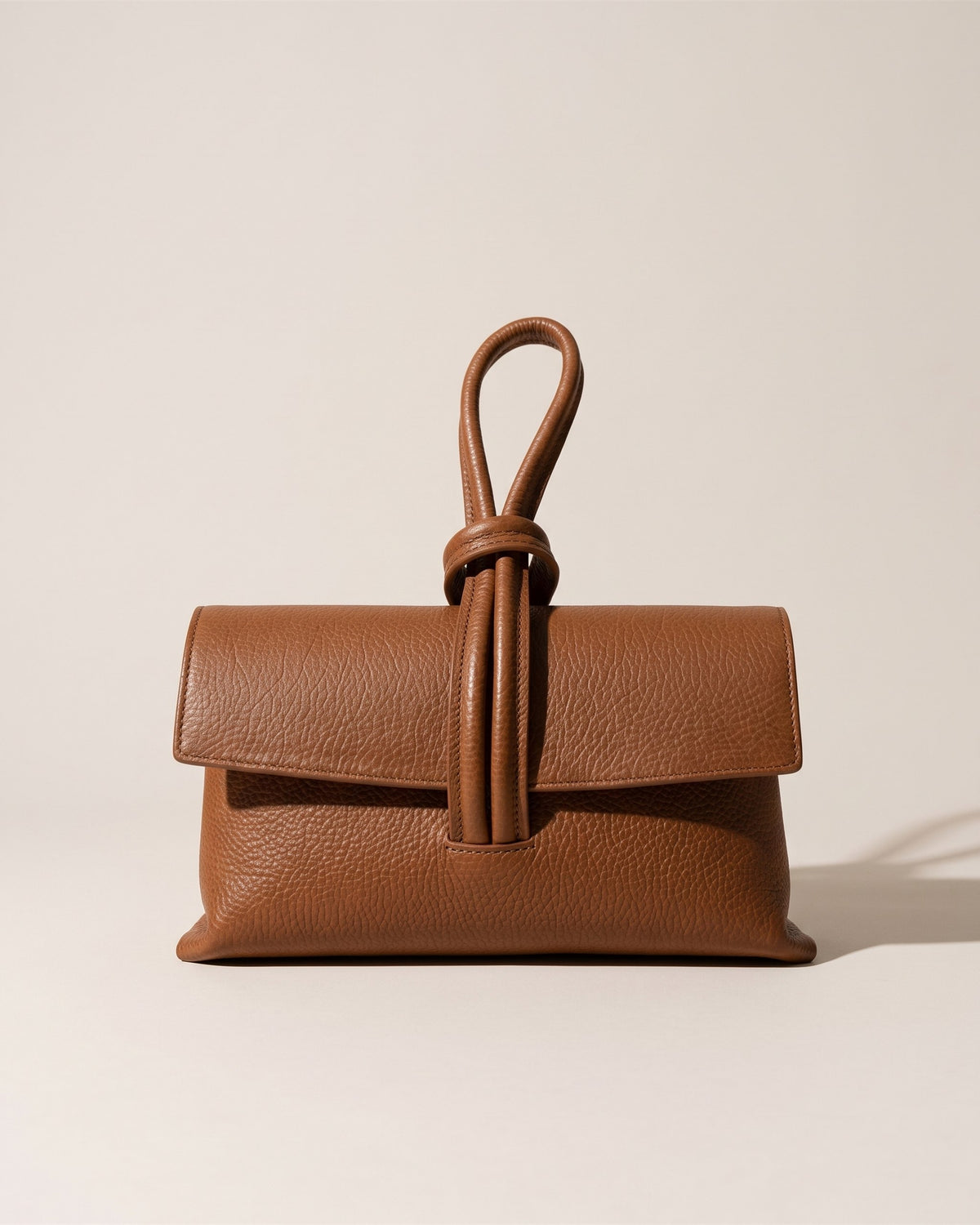 La pochette Siena en cuir grainé camel, rabat replié et anse boucle en cuir sur le dessus, vue de face sur fond pierre