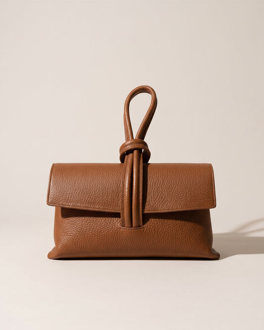 La pochette Siena en cuir grainé camel, rabat replié et anse boucle en cuir sur le dessus, vue de face sur fond pierre