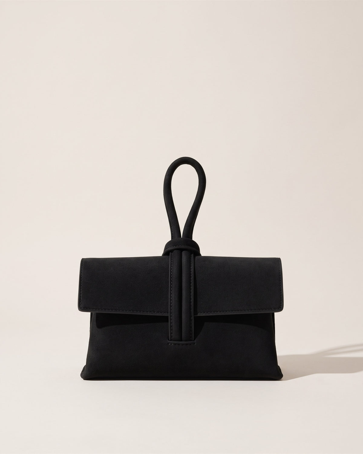 La pochette Capri en cuir suède noir, rabat replié et anse boucle en cuir sur le dessus, vue de face sur fond pierre