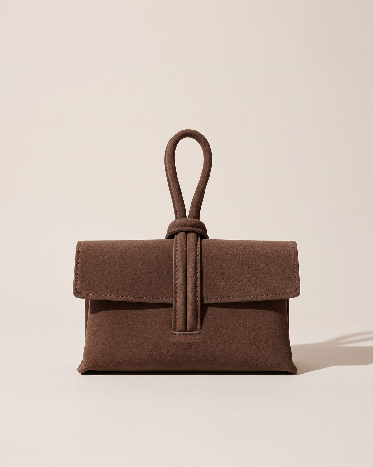 La pochette Capri en cuir suède chocolat, rabat replié et anse boucle en cuir sur le dessus, vue de face sur fond pierre